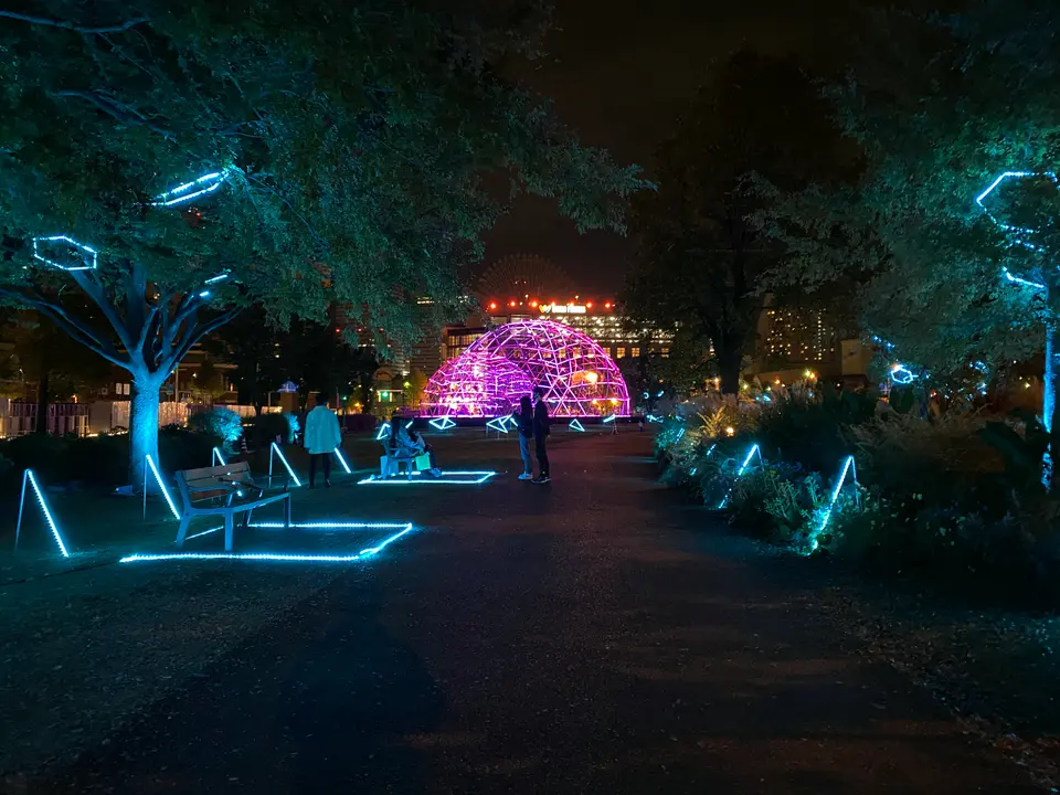 青い光に包まれた公園の遊歩道と、紫色に光る「CROSS DOME」の夜景