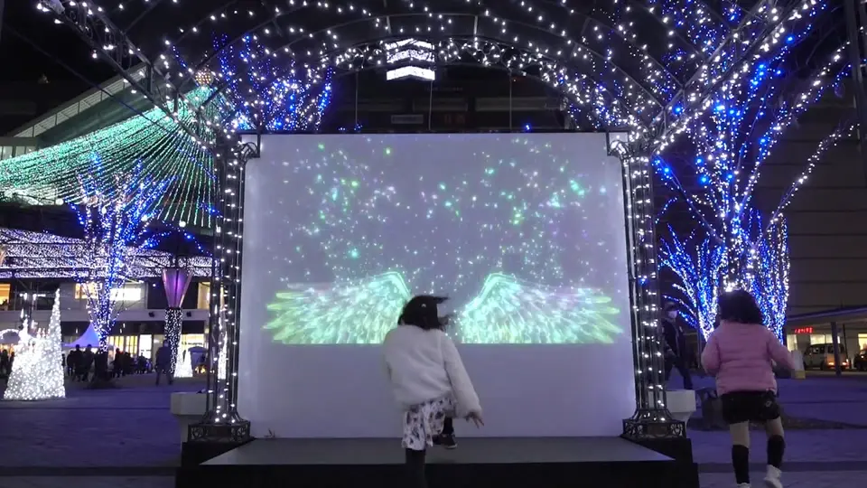 光のアーチに囲まれた夜の「インタラクティブ フォトスポット」。天使の羽が舞台に映し出される様子